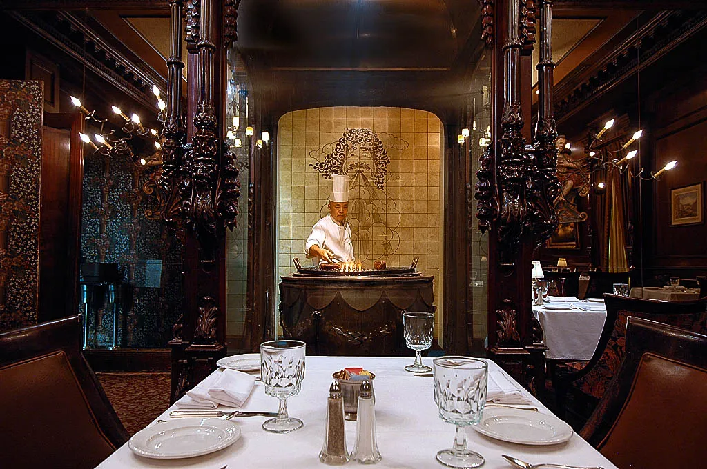 Chef preparing food in a gourmet restaurant kitchen, with an elegant dining area visible in the foreground.