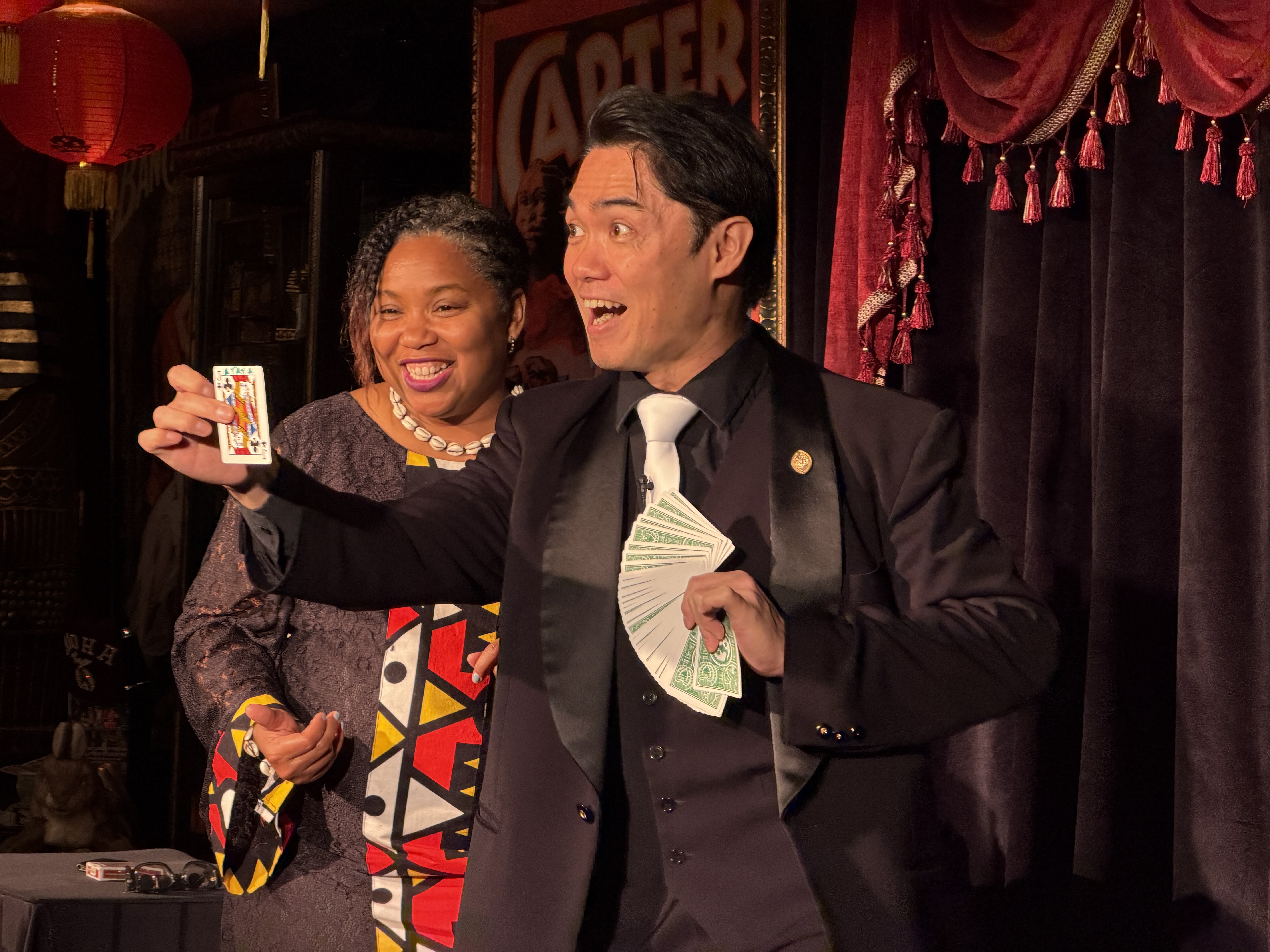 Magician performing a card trick with a guest, holding a deck of cards and money, while both share a moment of laughter during the performance.